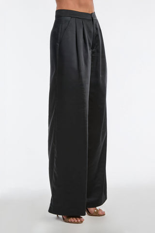 A person is wearing the Valentina Satin Pant in black, featuring high-waisted, wide-leg styling that creates a chic, flowy silhouette. Paired with tan open-toe heels, the satiny fabric shines against a plain white background.