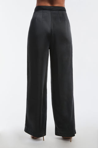 Someone is seen from behind wearing the Valentina Satin Pant in black, featuring high-waist and wide-leg styling. The smooth, glossy satin fabric highlights the subtle seam details against a plain white backdrop.