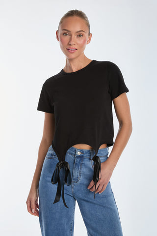 Wearing a Diana Bow T-Shirt in black featuring an asymmetric hem and satin bow-detail ribbon ties, a person pairs it with blue jeans against a plain white background, showcasing a pleasant expression.