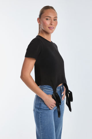 A woman wearing a black Diana Bow T-Shirt with side ties against a white background pairs it with Rosalie Jeans. Her hair is pulled back as she looks to the side, smiling gently, embodying casual elegance.