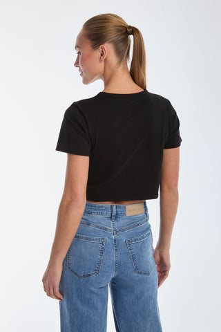 A person with a ponytail is wearing a black Diana Bow T-Shirt and Rosalie Jeans, standing with their back to the camera and looking sideways. The satin bow-detail adds charm against a plain light-colored background.