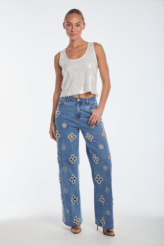 In a studio, a woman smiles with her hair tied back, wearing a white sequin sleeveless top and Kaia Pearl High Rise Jeans in medium blue. The jeans, high-waisted with beaded flower embroidery, complement her effortless style as she poses confidently in heels.