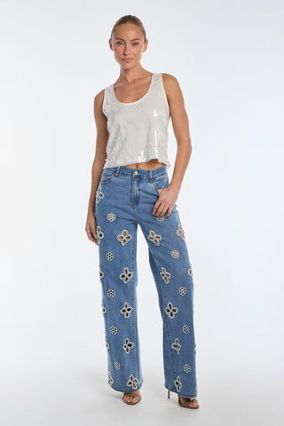 A woman exudes confidence against a white backdrop, showcasing a shiny sleeveless top and Kaia Pearl High Rise Jeans in Medium Blue. The jeans flaunt floral cut-outs and embroidered beaded flowers on luxurious stretch denim. Her elegant high heels complement her tied-back hair perfectly.