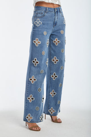 The woman wears Kaia Pearl High Rise Jeans in medium blue, showcasing wide-leg denim with embroidered beaded flowers, paired elegantly with a white top and open-toe heels against a plain background.