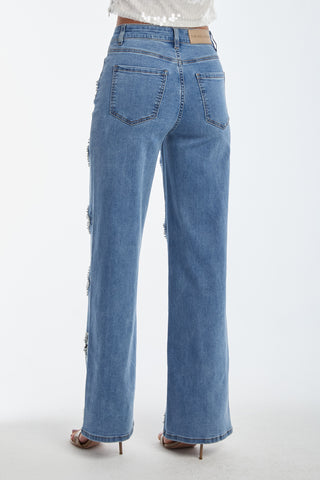 Rear view of a person in medium blue Kaia Pearl High Rise Jeans with frayed sides. These straight-leg jeans feature back pockets, a branded leather patch on the waistband, and embroidered beaded flowers, paired with nude heels.