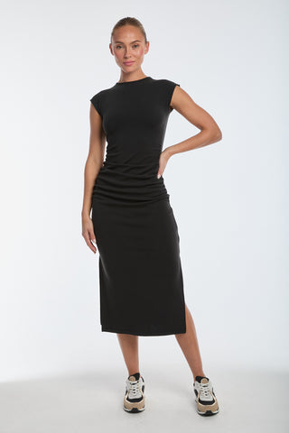 A person confidently wears the Daphne Dress - Black with side ruching, paired with black-and-white sneakers. The plain white background highlights the elegant outfit made from ultra-soft heathered modal.