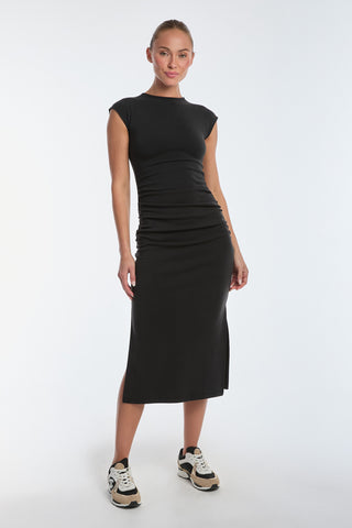 A person in a fitted Daphne Dress - Black with side ruching and a slit stands on a plain white background, pairing it with black and white sneakers. The ultra-soft heathered modal fabric adds to the look as their hair is pulled back and they smile slightly.