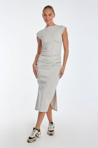 Wearing a form-fitting Daphne Dress - Heather Grey, made of ultra-soft heathered modal with side ruching and a slit, a person stands confidently against a plain background. They pair it with black and white sneakers, posing with one hand on their hip.