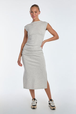 A woman confidently poses in a Daphne Dress - Heather Grey, crafted from ultra-soft heathered modal with elegant side ruching. She stylishly pairs it with black and white sneakers against a plain white background.