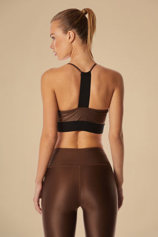 A woman with a ponytail showcases the Liquid Studio Bra - Cafe, featuring a sporty elastic back and scoop neckline, paired with matching high-waisted leggings. She faces away from the camera against a beige backdrop, emphasizing the outfit's fit.