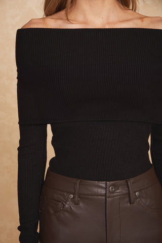 A person is wearing an Amora Ribbed Knit Top - Black from Noli Yoga, paired with high-waisted brown leather pants. The image captures from the shoulders to the upper thighs, with the head not visible. The background is a beige-colored wall.