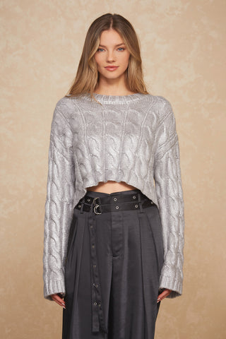 A woman with long blonde hair wearing a Metallic Cropped Sweater - Silver by Noli Yoga and high-waisted dark gray pants with belt details is standing against a light textured background, looking directly at the camera and showcasing her chic fall wardrobe.