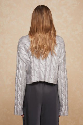 A woman with long, wavy, chestnut hair is seen from behind, wearing the Metallic Cropped Sweater - Silver by Noli Yoga and black pants. The background is a textured, beige wall.