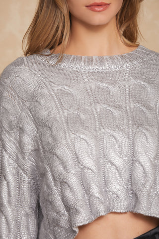 A close-up of a person wearing the Metallic Cropped Sweater - Silver by Noli Yoga, featuring its eye-catching cable-knit design with a shimmering finish. The sweater's shiny texture and intricately detailed knit pattern make it an ideal addition to any Fall wardrobe. The person has long, light brown hair, and only their neck, lower face, and upper torso are visible.