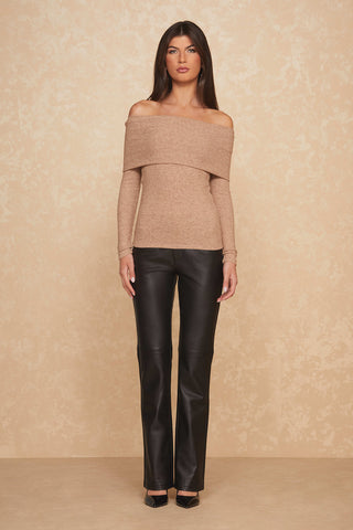 A woman stands against a beige textured background, wearing an ultra-soft off-the-shoulder Amora Top in Praline and black leather pants. She has long, straight brown hair and dons black pointed-toe shoes. Her arms are relaxed by her sides.