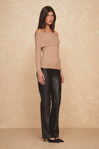 A woman stands against a beige textured background, wearing an ultra-soft, off-the-shoulder Amora Top in Praline and black leather pants. She has long, dark hair and is wearing black high-heeled shoes. She poses with her hands at her sides, looking at the camera.