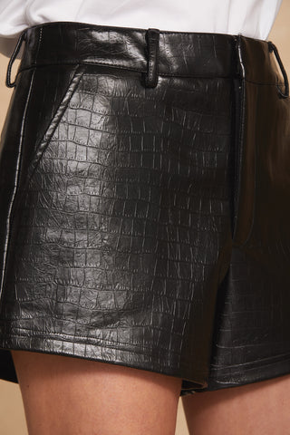 Close-up of a person wearing the Jaclyn Leather Short in black by Noli Yoga, exuding an on-trend look. The faux croco embossed leather shorts feature belt loops and are paired with a tucked-in white top. The skin of the person's legs is partially visible.