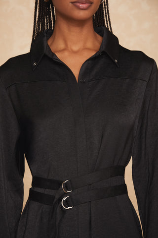 A person with braided hair is wearing the Simone Double Belted Satin Dress by Noli Yoga. The long-sleeved black dress, made from textured satin, features a collar, buttons down the front, and a double belt with metal rings at the waist. The background is light beige. Only the person's torso and lower face are visible.