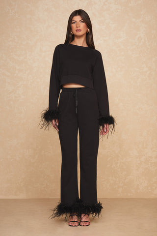 A glamorous woman stands against a beige textured background, dressed in Noli Yoga's Rada Feather Trim Jogger - Black, an elegant black long-sleeve top and matching black pants adorned with feather trim on the sleeves and pant legs. She has long brown hair, black high-heeled sandals, and gold hoop earrings.