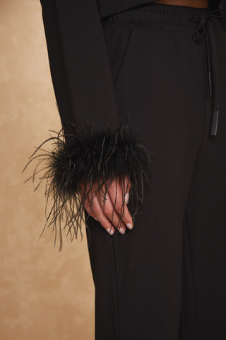 Close-up of a person wearing black pants and a matching dark Noli Yoga Rada Feather Trim Crewneck. The attire appears elegant and formal, with the unique touch of feathered cuffs adding glamorous texture to the wrist area. The background is a subtle beige.