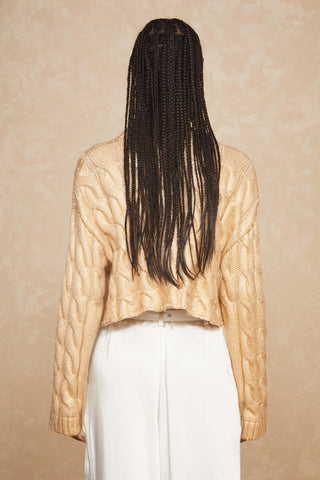 A person with long braided hair is seen from the back, wearing a gold Metallic Cropped Sweater by Noli Yoga and white high-waisted pants; the perfect addition to any Fall wardrobe. The background is a textured light beige, complementing the outfit beautifully.