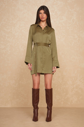 A person with long hair is wearing the Simone Double Belted Satin Dress in Olive by Noli Yoga, paired with knee-high brown boots. The figure-flattering ensemble elegantly complements the beige, textured background.