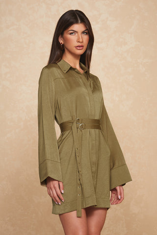 A woman stands against a textured beige background, wearing the figure-flattering Simone Double Belted Satin Dress in olive from Noli Yoga. The elegant dress features a collar and slightly flared long sleeves. She has long, dark hair and gazes directly at the camera with a neutral expression.