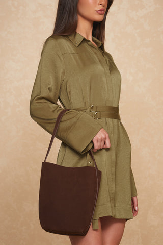 A person wearing a light olive green long-sleeve romper with a tied waist is holding the Paris Bag in Java Suede by its shoulder strap. The background is a soft, neutral beige color with a slight texture.