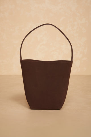 A minimalist, dark brown Paris Bag - Java Suede bucket bag with a single strap, displayed upright against a muted beige background. The bag features a simple, sleek design with a slightly tapered shape toward the base.