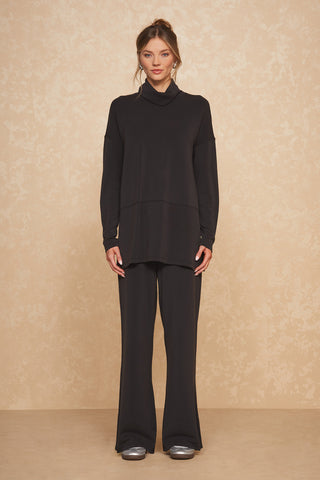 A person stands against a beige textured background, wearing a loose black turtleneck sweater paired with the Daphne Wide Leg Pant - Black from Noli Yoga. They have light-colored sneakers and their hair is tied up in a casual bun. The soft modal blend fabric of the outfit exudes luxurious comfort, and their hands are relaxed by their sides.