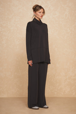 A person stands against a beige textured background, modeling the black Daphne Top from Noli Yoga paired with wide-leg black pants. They wear silver shoes and have their hair styled up, with long earrings visible. The attire gives a modern, minimalist aesthetic and exudes luxurious comfort.