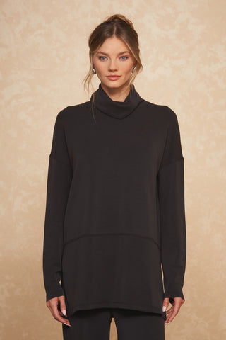 A woman stands against a beige textured background, wearing the Daphne Top in black - a loose-fitting, long-sleeved top with a high collar made from a soft modal blend. Her hair is styled in a loose updo and she has a neutral expression on her face, exuding luxurious comfort.