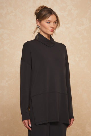 A woman stands against a beige textured background, wearing the Daphne Top in black, which features long sleeves and a high neckline. Crafted from a soft modal blend, her attire exudes luxurious comfort. She has her hair styled in an elegant updo and is adorned with subtle jewelry, including earrings. She gazes slightly to the side with a calm expression.