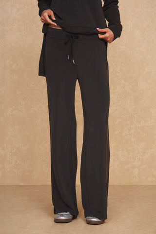 A person wearing the Daphne Pant in black, made from a soft modal blend, and silver shoes stands against a beige, textured background. Only the lower torso and legs are visible. The person is adjusting the waistband of these luxurious comfort pants with their hands.