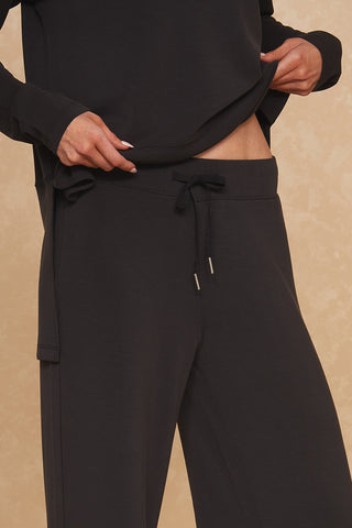 A person wearing a black long-sleeve Daphne Top and Noli Yoga's black Daphne Wide Leg Pant, characterized by its drawstring waistband, is seen holding the bottom of the shirt with one hand, pulling it slightly upward. The background features a textured beige wall that accentuates the luxurious comfort of the soft modal blend fabric.