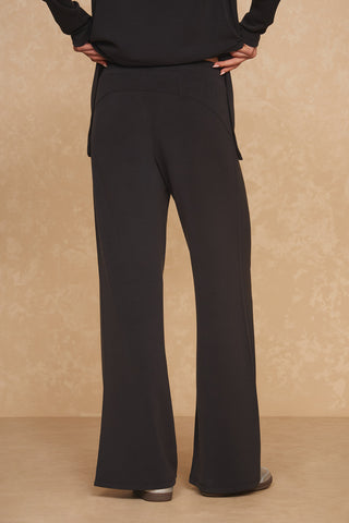 A person stands with hands on hips, facing away from the camera, wearing black Daphne Wide Leg Pants and a black long-sleeve top made of soft modal blend, both from Noli Yoga. The luxurious comfort is complemented by the textured beige wall in the background.