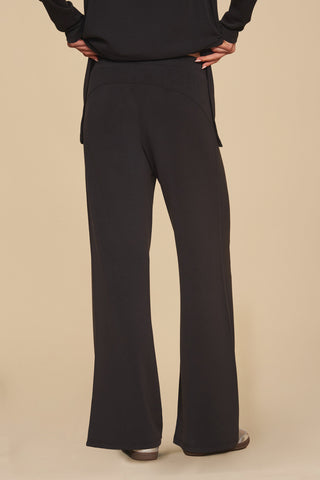 A person wearing a pair of Daphne Pant - Black and a Daphne Top stands against a beige background. The view from behind highlights the relaxed fit and wide legs of the pants made from a soft modal blend. The ensemble is complemented by shoes with a slight heel, enhancing its luxurious comfort.