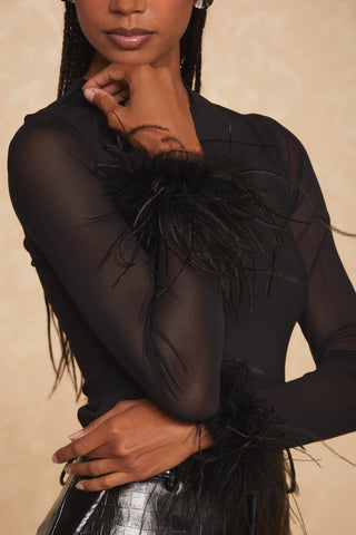 The individual is donning the Zinna Sheer Feather Top in black by Noli Yoga, featuring sheer long sleeves decorated with an ostrich feather trim at the cuffs. The image is cropped from the chest down, revealing their left hand resting on their right arm against a neutral beige background, highlighting the elegance of this sophisticated statement piece.