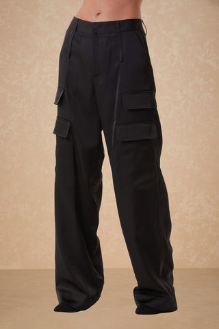 A person is wearing the Milan Pant in black, featuring high-waisted, wide-leg cargo trousers with multiple pockets. The background is a beige wall with a subtle textured pattern. The person’s upper body is not visible in the image.