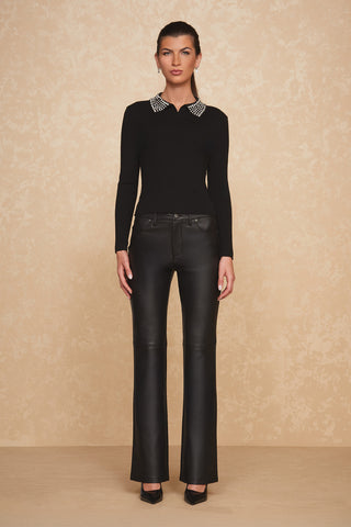 A person stands against a beige background wearing the Crystal Polo Sweater - Black, a sleek black long-sleeve top with crystal beaded embellishments on the white collar, paired with black leather pants and black pointed heels. The person has their hands resting by their sides and is facing forward.