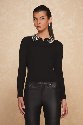 A woman dressed in the Crystal Polo Sweater - Black, a long-sleeve top adorned with crystal beaded embellishments on the collar, pairs it with black leather pants. She stands against a beige textured background, her long dark hair tied back and accessorized with small earrings. She poses confidently, one hand resting by her side.