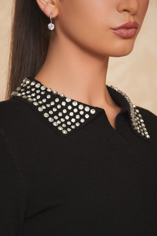 A woman with long dark hair is shown from the lower lip down, wearing drop earrings and the Crystal Polo Sweater in black, which features a bedazzled collar adorned with numerous crystal beaded embellishments. The background is a light beige color.