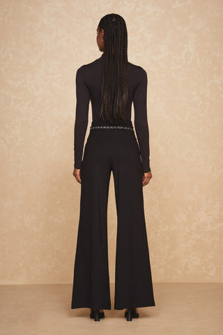 A person with long braided hair stands with their back to the camera. They are wearing a black, long-sleeved top paired with the Ava Pant in black, along with black heeled shoes. The background features a beige textured wall.