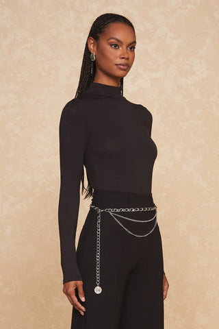 A person stands against a beige background, wearing a long-sleeve black turtleneck top and black pants. They have braided hair and wear dangling earrings. A trendy silver chain belt, identified as the Jane Belt - Silver, with a circular pendant accents the outfit, adding a touch of elegance.