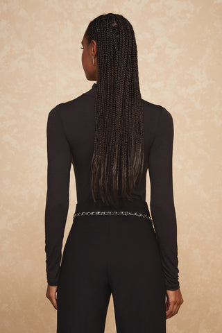 A person with long braided hair stands facing away, dressed in a fitted black long-sleeve top and pants. A trendy accessory—a thin silver chain belt, known as the Jane Belt—perfectly accentuates the outfit against the textured light brown backdrop.