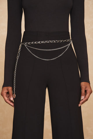 The image features a person wearing a black long-sleeve top and black high-waisted pants, accessorized with the stylish Jane Belt in silver, draped elegantly around the waist. The background is a neutral beige.