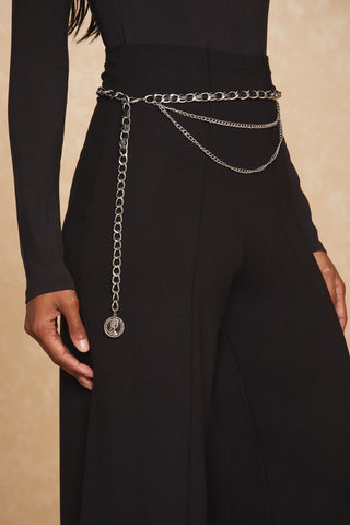 A person wearing the Ava Pant - Black and a long-sleeve black top is shown. The wide-leg pant flows elegantly, complemented by a silver chain belt with layered chains and a hanging pendant wrapped around their sculpted waist. The background is a soft, light beige color.