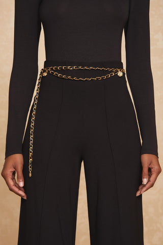 A person wearing a black long-sleeve top and high-waisted black pants accessorized with the stylish Jane Belt in gold, which wraps around the waist and ends with a small gold charm. The person's arms are by their sides, and the background is textured in a warm, neutral tone.