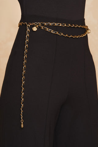 A close-up view of the Jane Belt - Gold, featuring a Medusa emblem, wrapped around the waist of a person wearing high-waisted black pants. The chain drapes elegantly, adding a stylish touch to the trendy accessory and sleek outfit. The background is a neutral beige color.