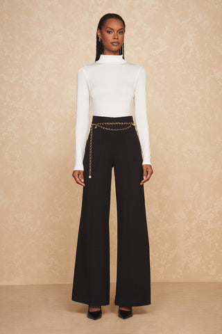 A person stands against a neutral beige background, wearing a white long-sleeve turtleneck top paired with the Ava Pant - Black. A gold chain belt accentuates the sculpted waist of the high-waisted, wide-leg trousers, and they are wearing black pointed-toe heels.